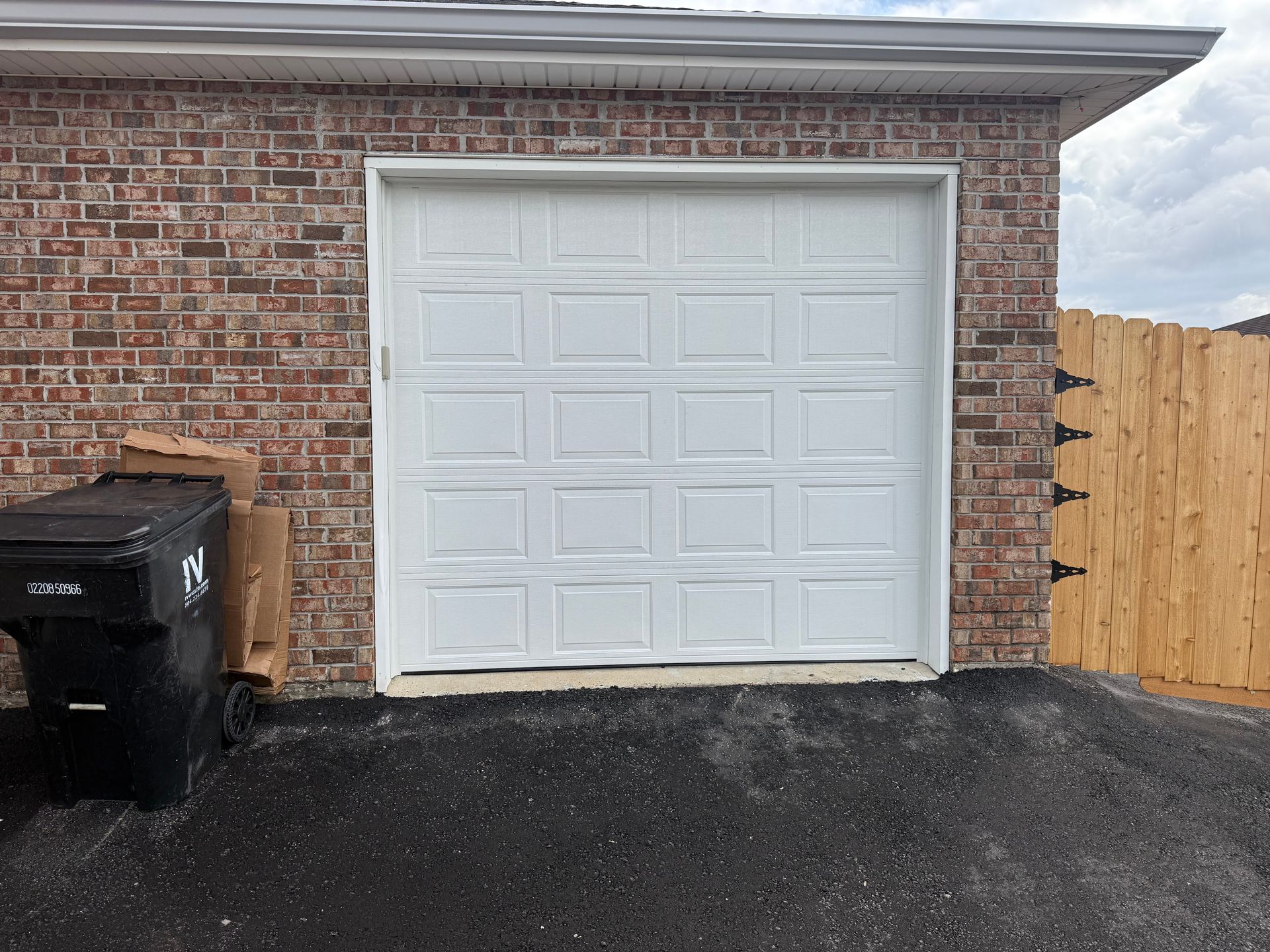 White garage door framed by brick and asphalt, trash can to the left, wooden fence to the right.
