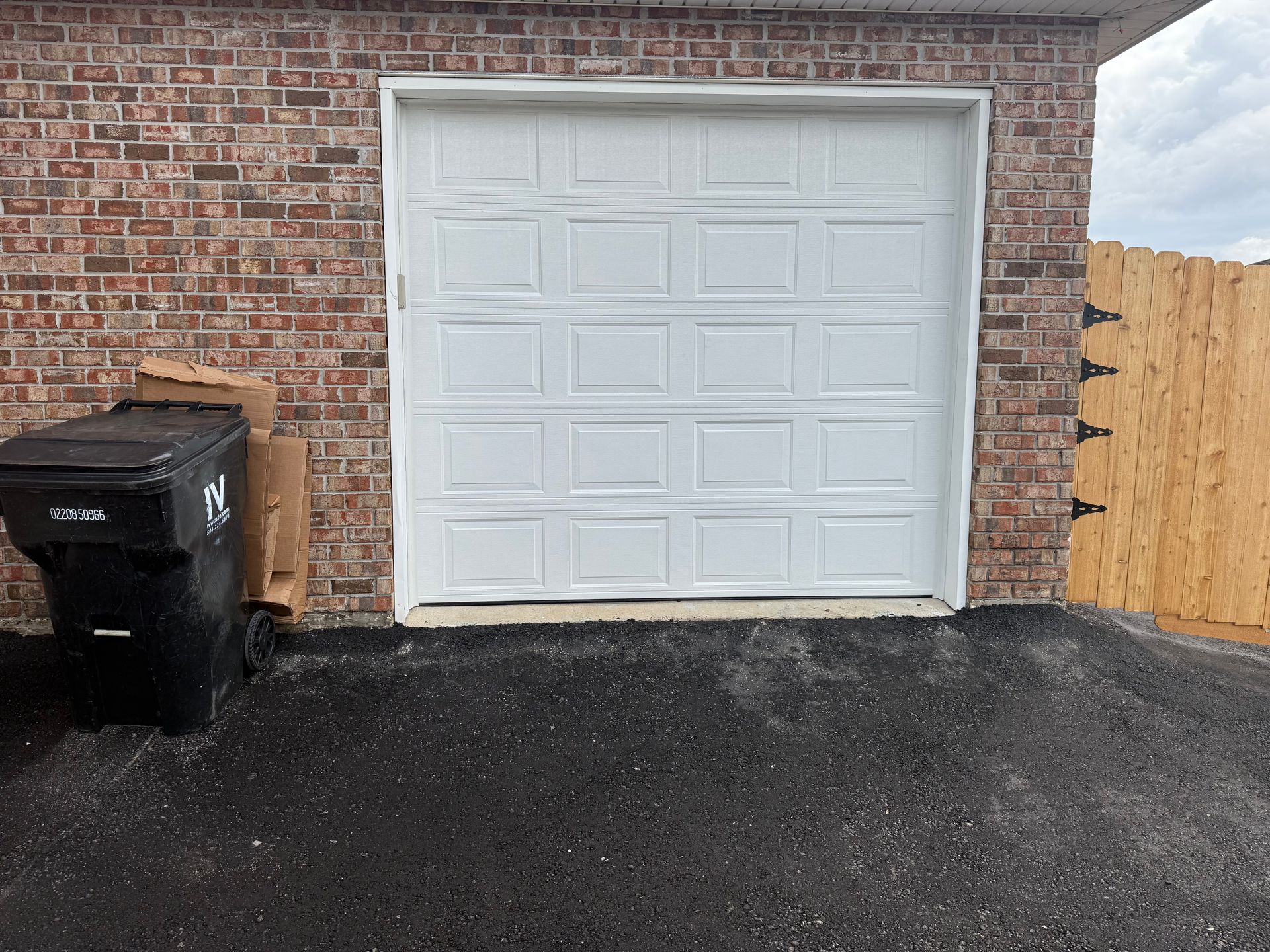 White garage door in brick building with trash can to the left and wooden fence to the right.