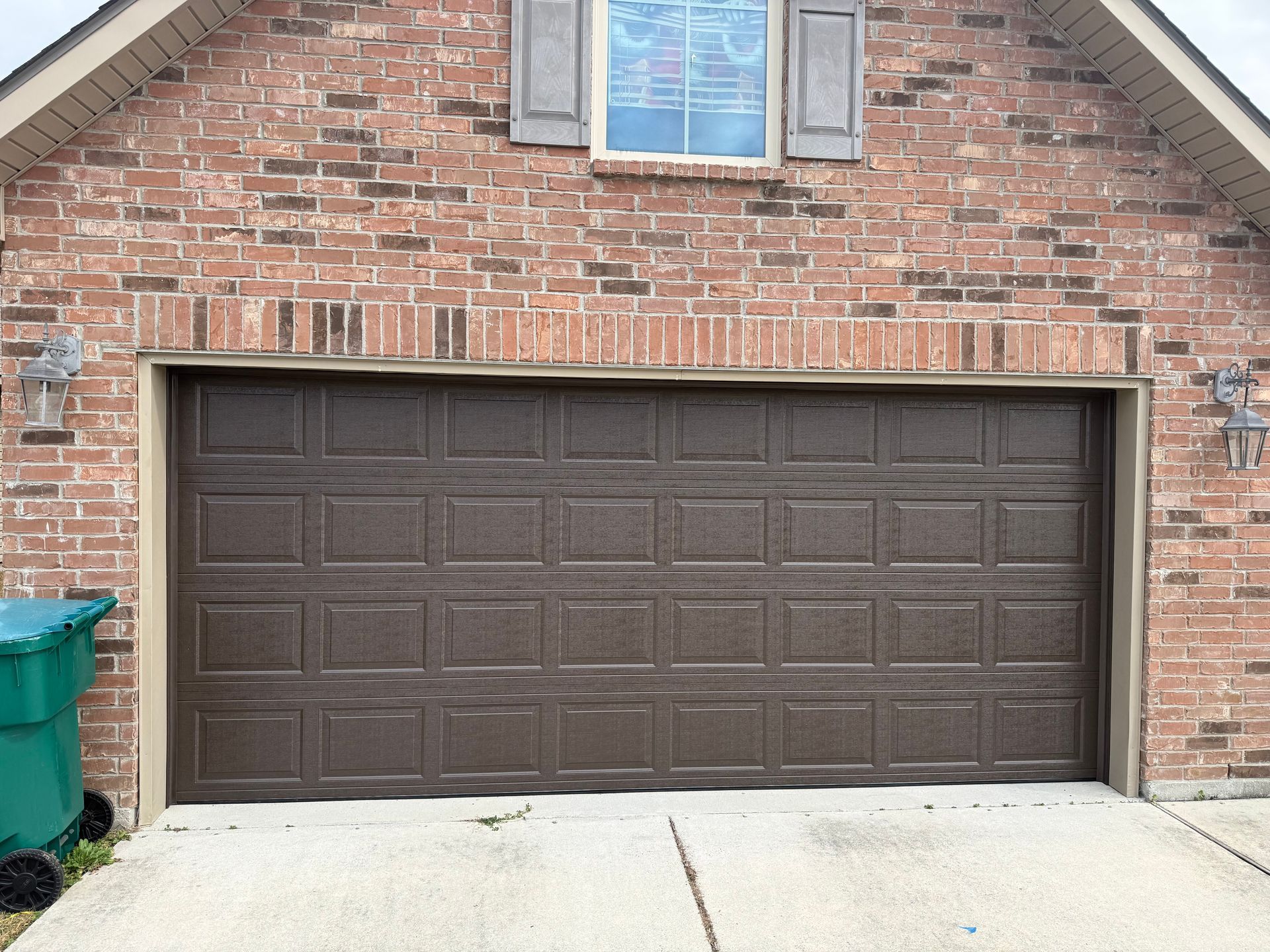 Brown garage door on a brick house with a window above. A green trash can sits beside it.