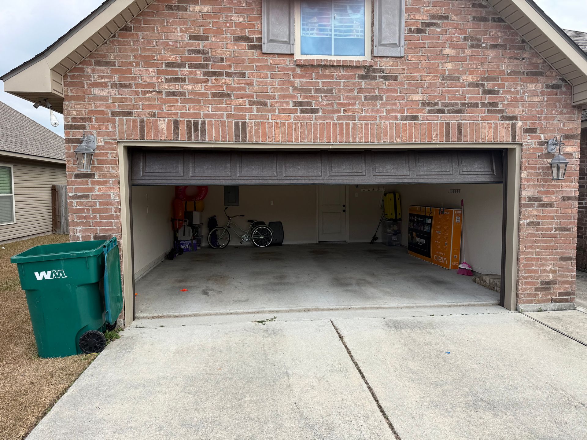 Exterior view of a brick house with an open garage. A green trash bin sits to the left of the garage door.