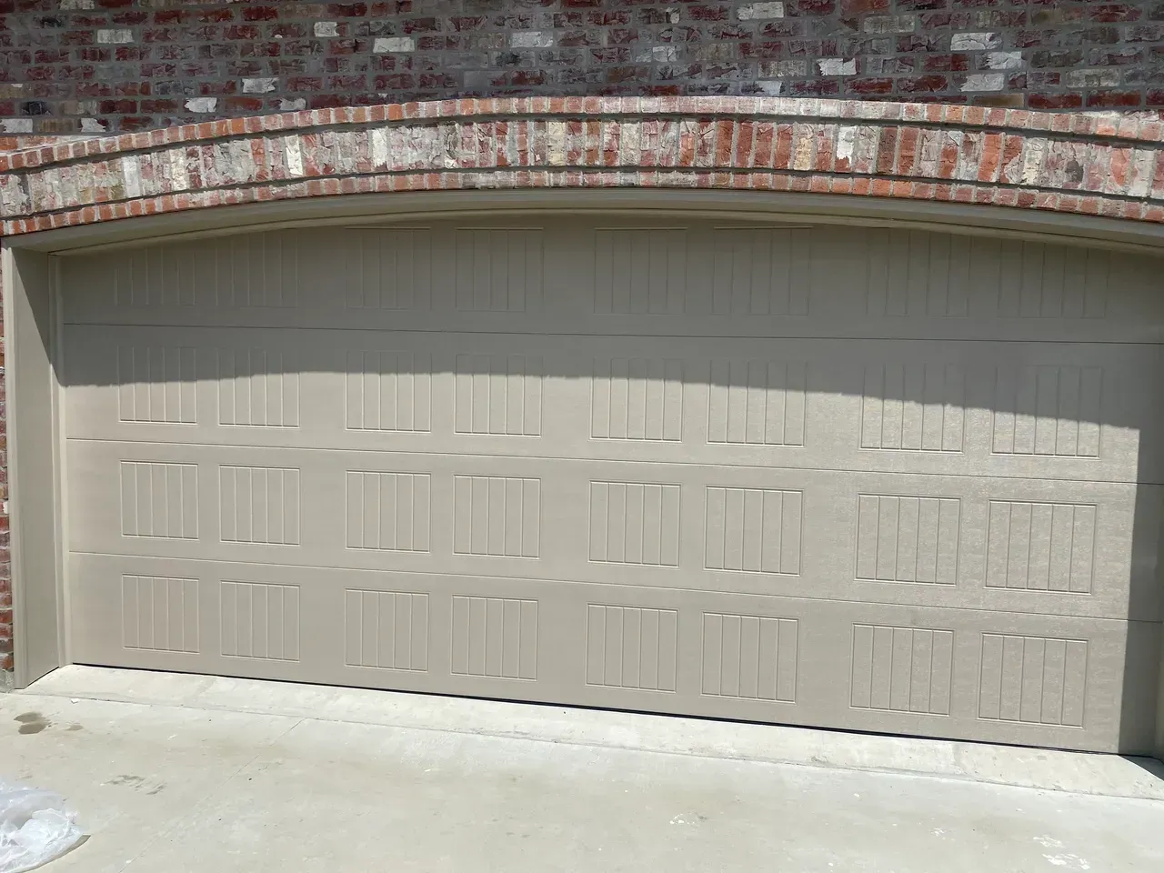Tan garage door with brick archway above, set in a concrete driveway.