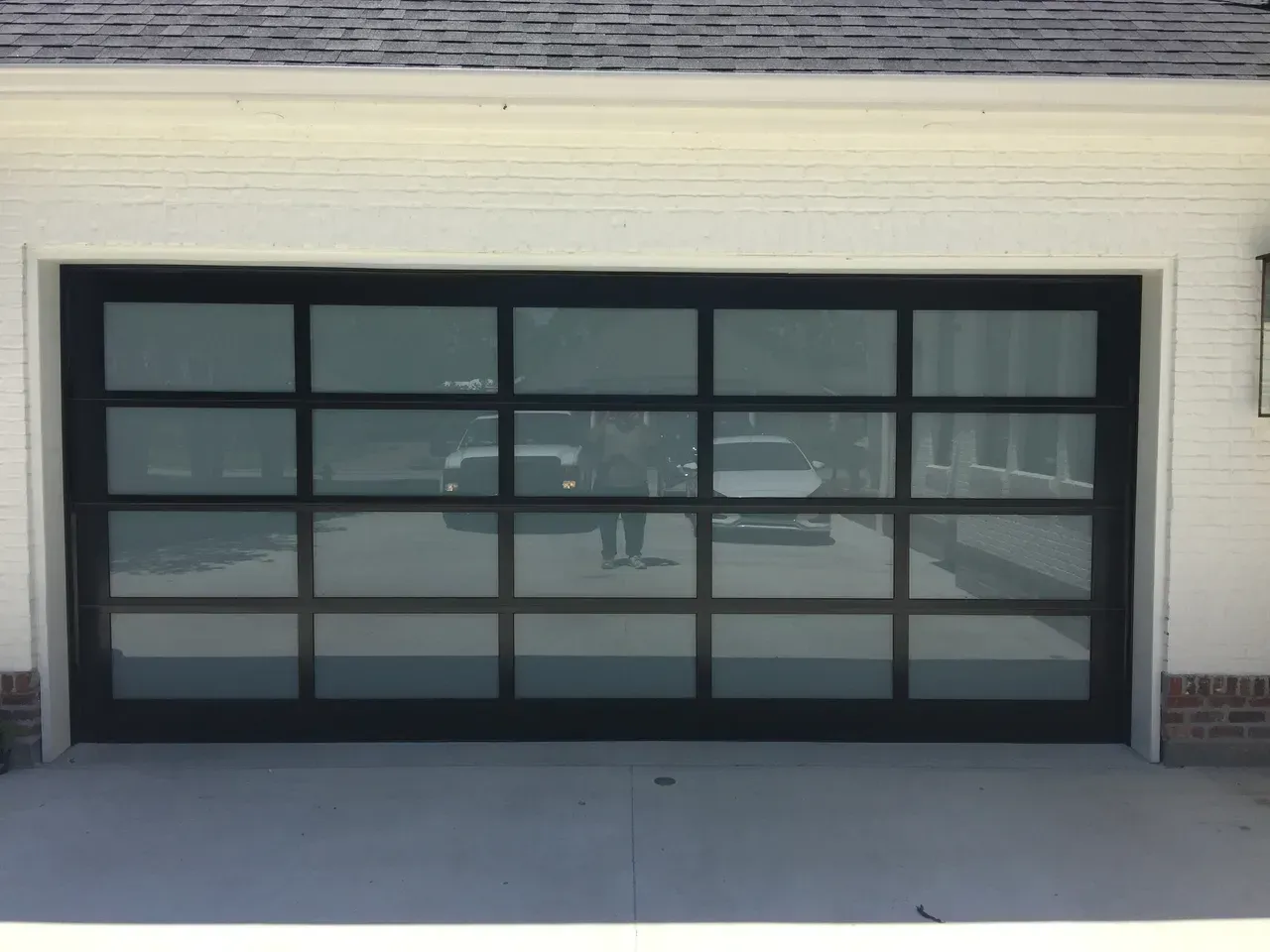 Black-framed glass garage door reflecting cars and person on a concrete driveway, set in a white building.