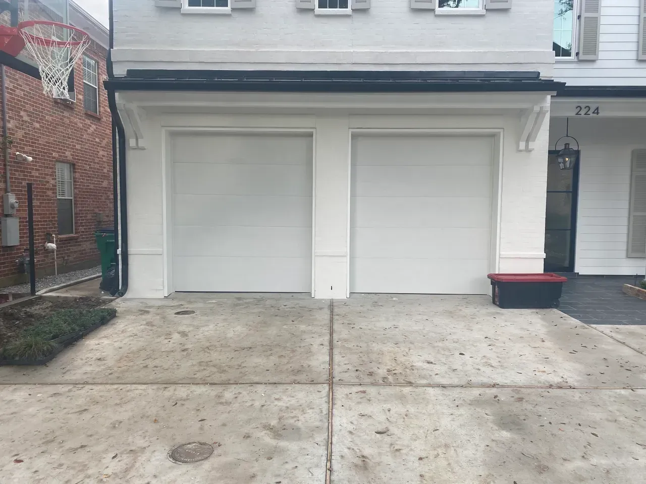 Two white garage doors on a light colored building; basketball hoop and red brick building on the left.