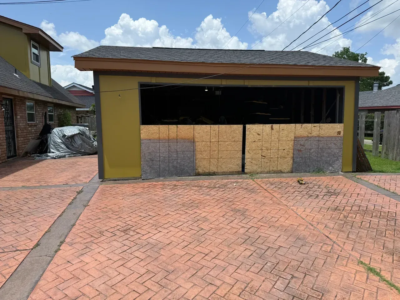 Garage with plywood covering the opening, fronted by a brick-paved driveway.