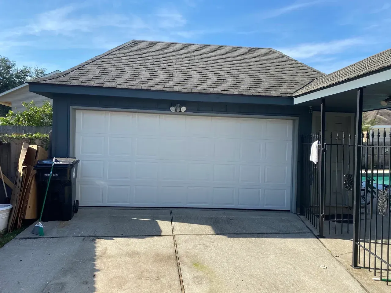 Garage with white door, gray roof, blue walls, and black trash can on concrete driveway.