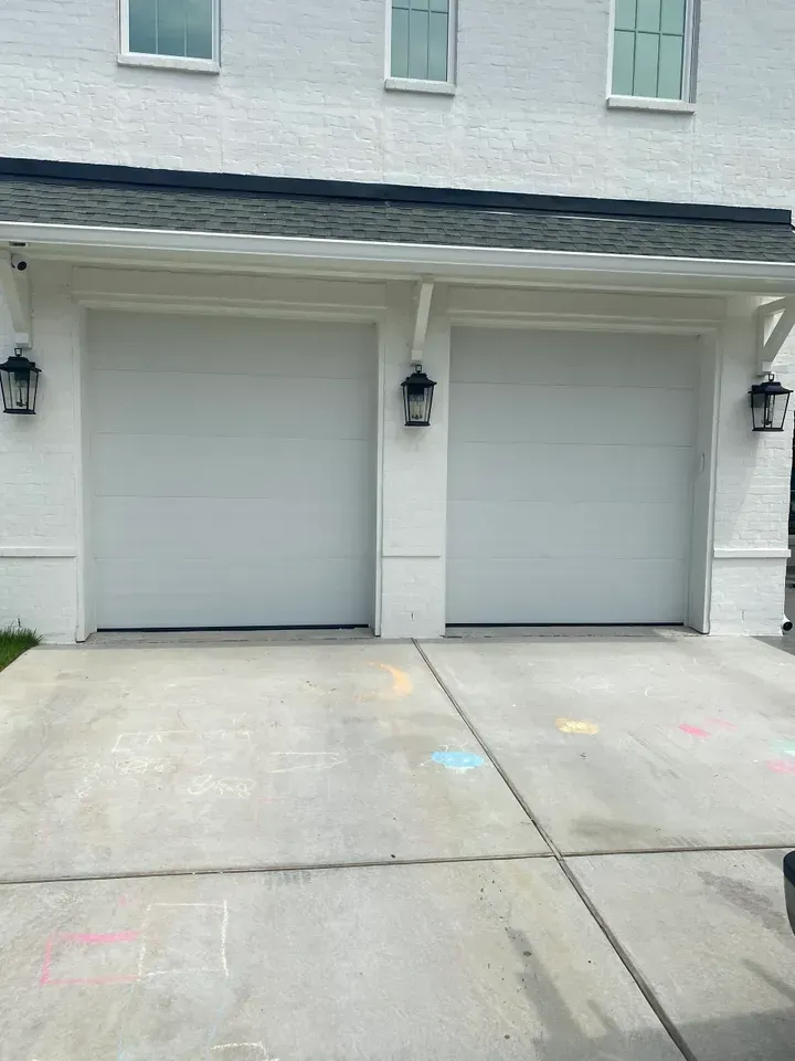 Two white garage doors on a white brick building with black light fixtures, concrete driveway with chalk drawings.
