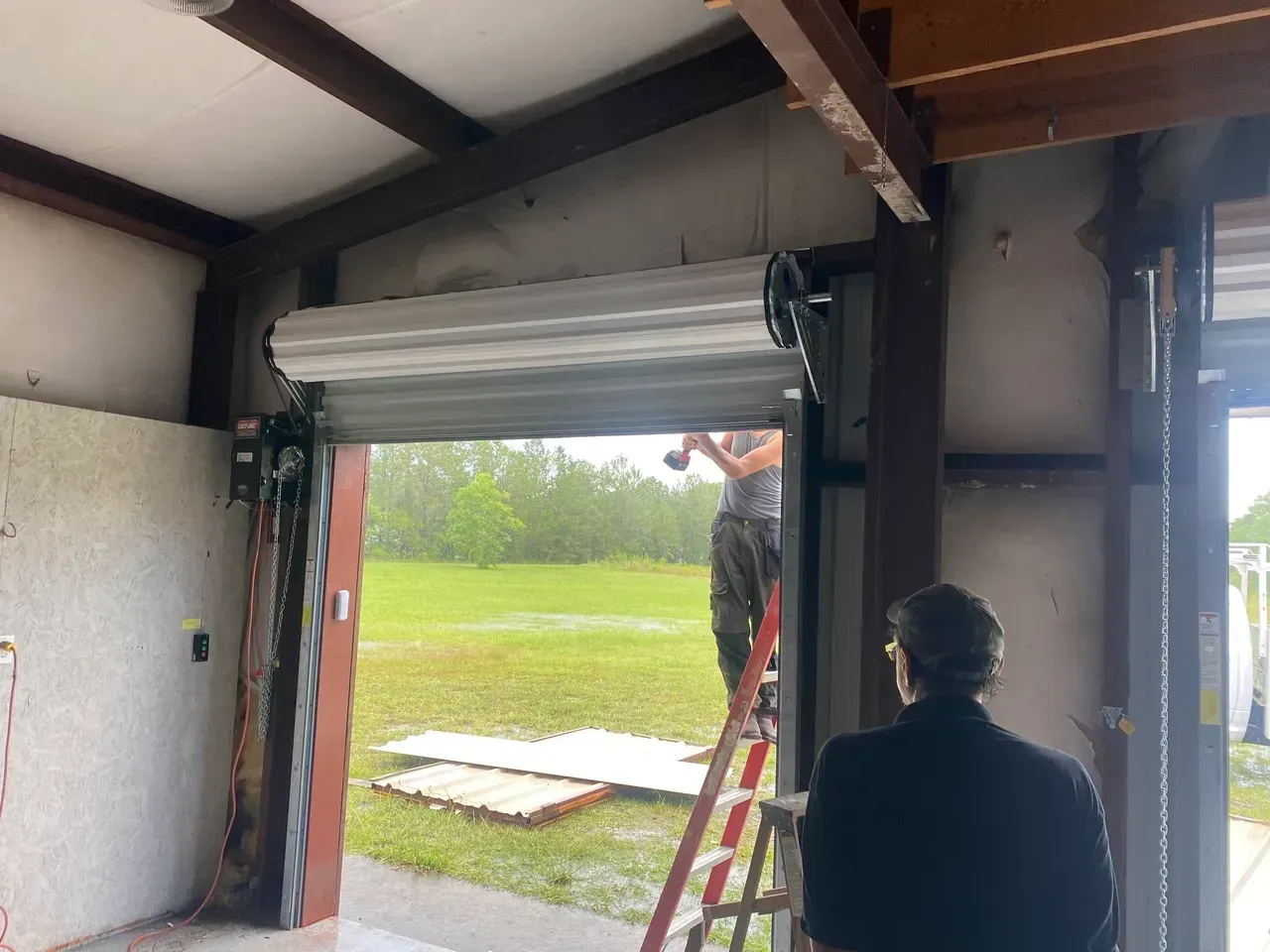Rolling metal security door partially open. Man on ladder, another person watches. Rainy outdoor view.