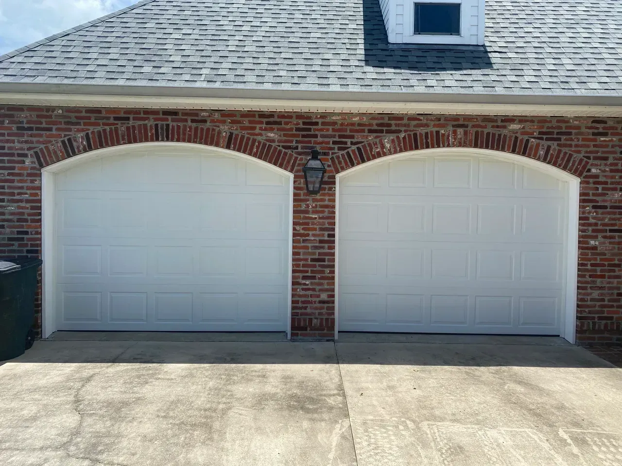 Two white garage doors under brick arches on a brick building with a gray roof and dormer.