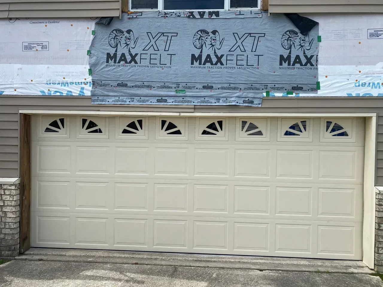 Beige garage door with arched glass windows above. Exterior siding and protective wrap visible above.