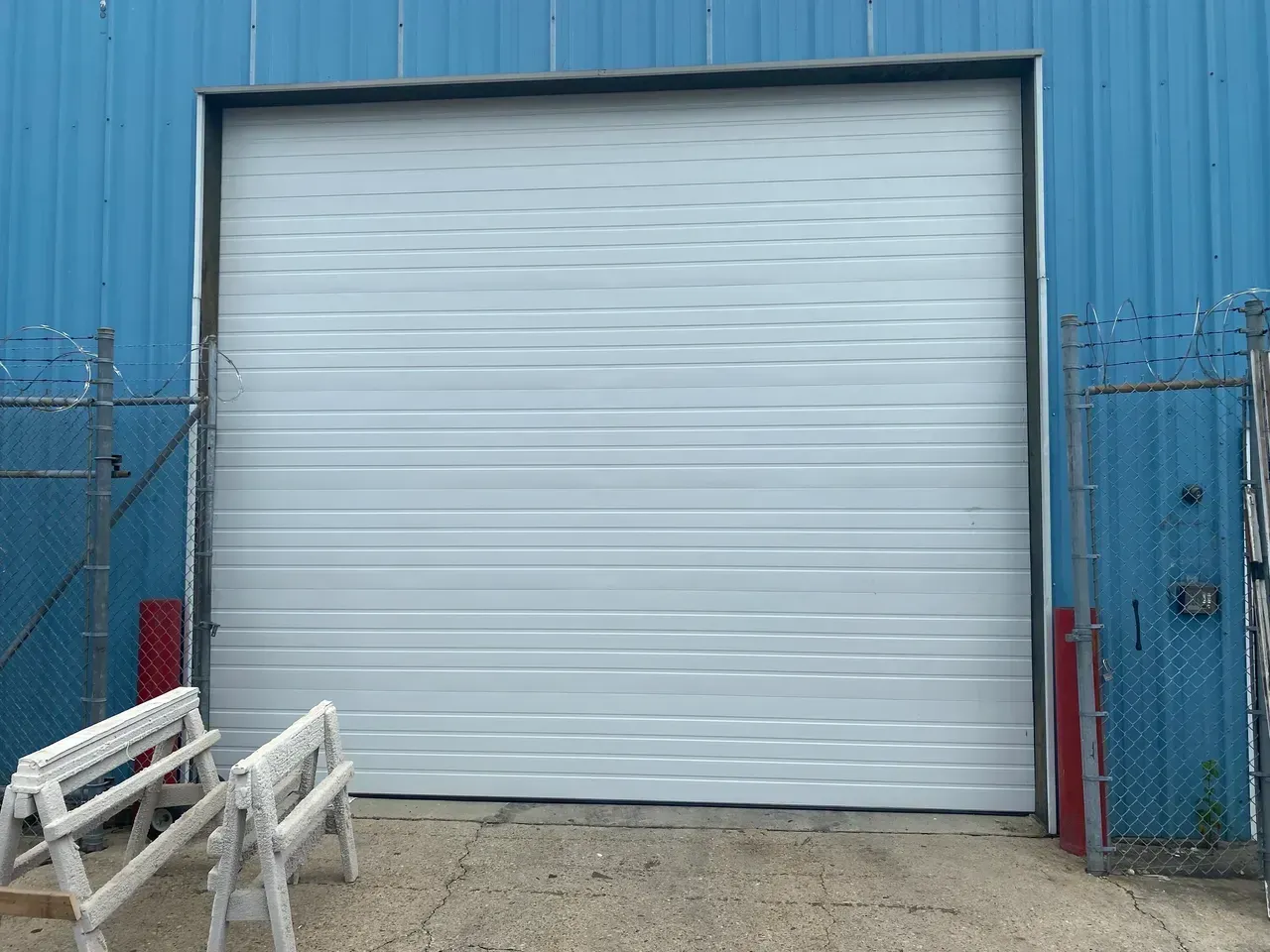 White closed overhead garage door on a blue building. Two saw horses and a chain link fence are nearby.