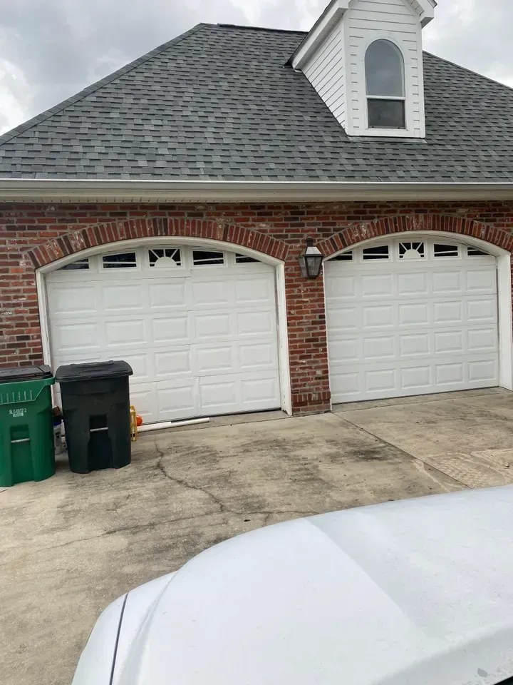 Two white garage doors on a brick house with a dormer and trash cans in front.