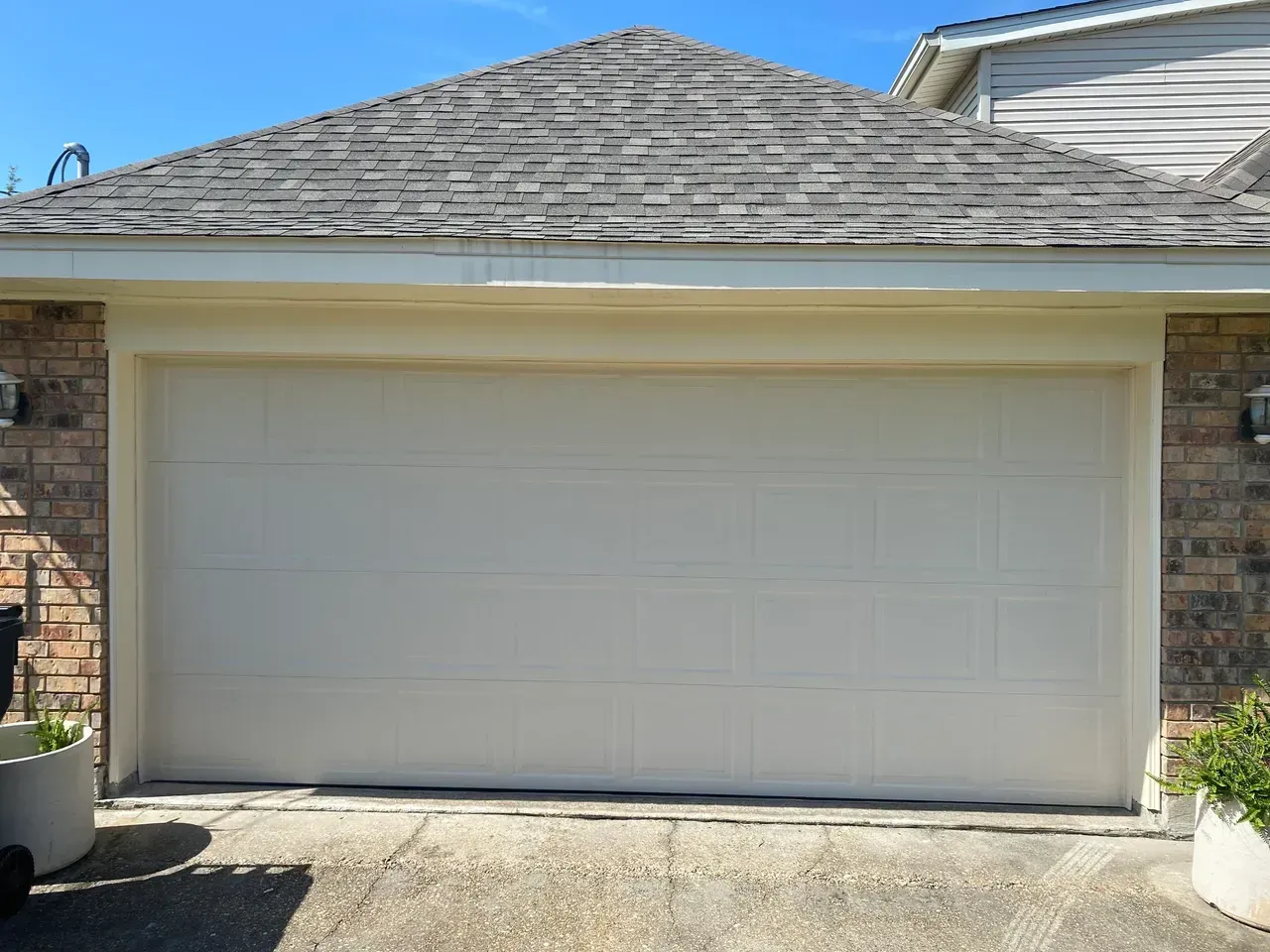 Tan garage door with a shingled roof, set against a brick wall.