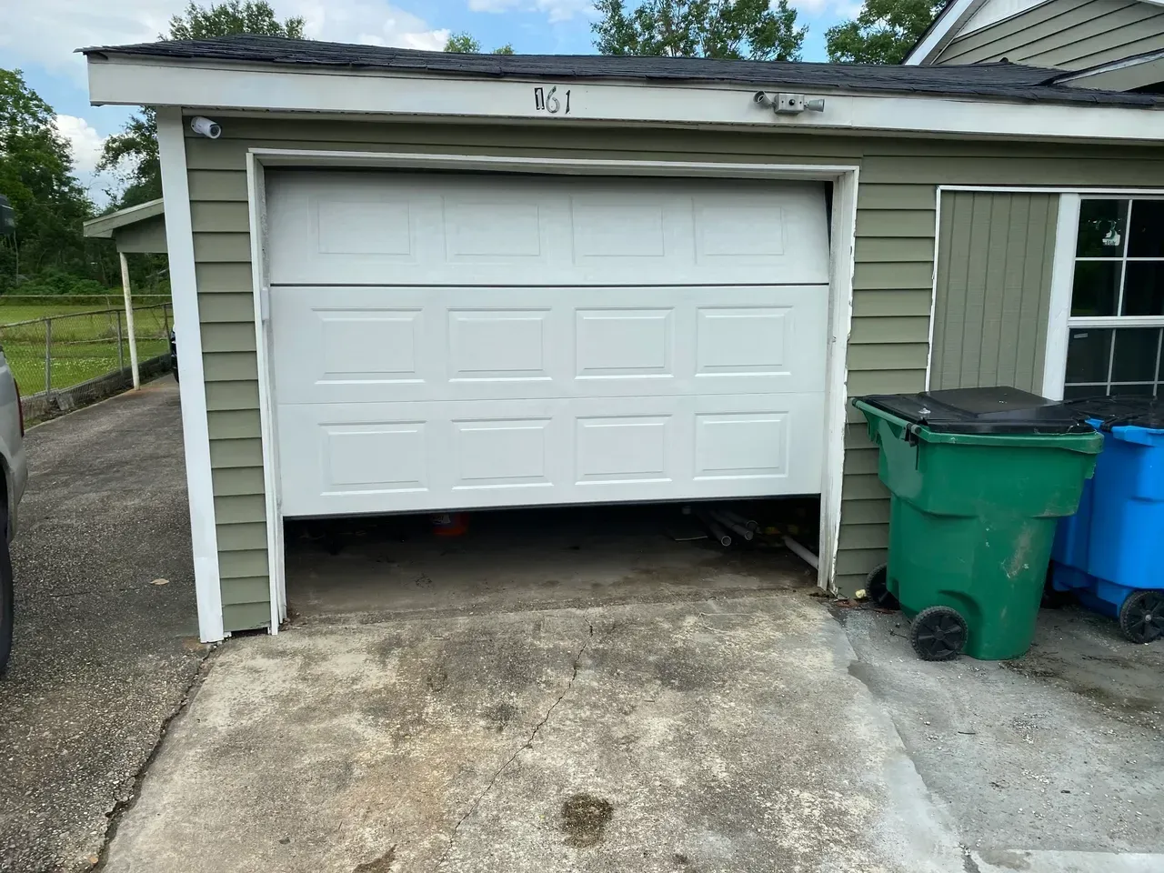 Garage door partially open; green siding, green and blue trash bins, concrete driveway.