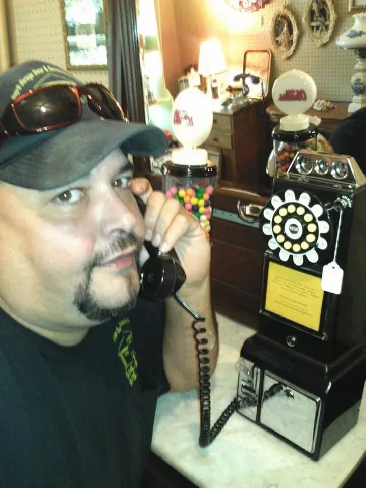Man holding a vintage black payphone receiver in an antique store; looking at the camera.
