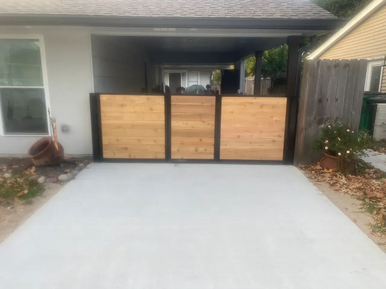Wooden gate with black frame, spanning a concrete driveway, enclosed by a building and a fence.