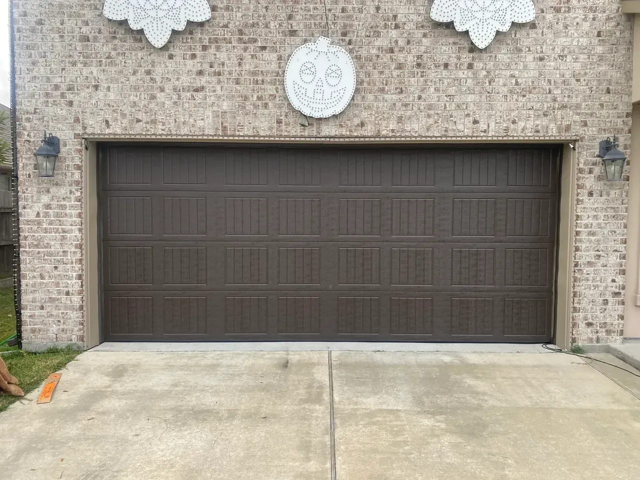 Brown garage door on a brick building with a concrete driveway. Decorative white ornaments above door.