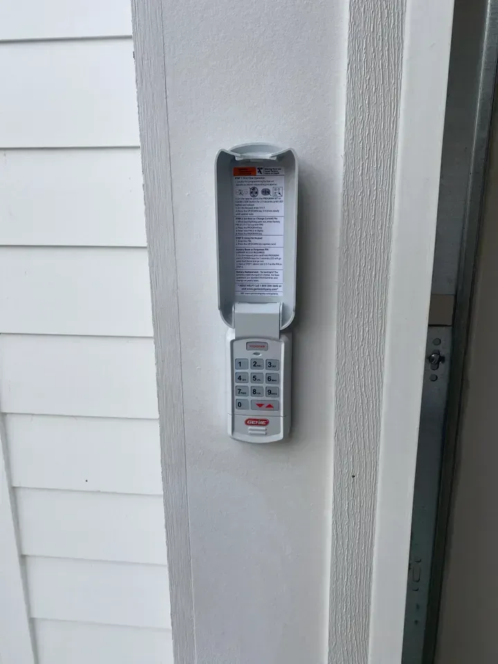 White garage door keypad mounted on a white door frame; keypad is open, showing a numbered keypad.