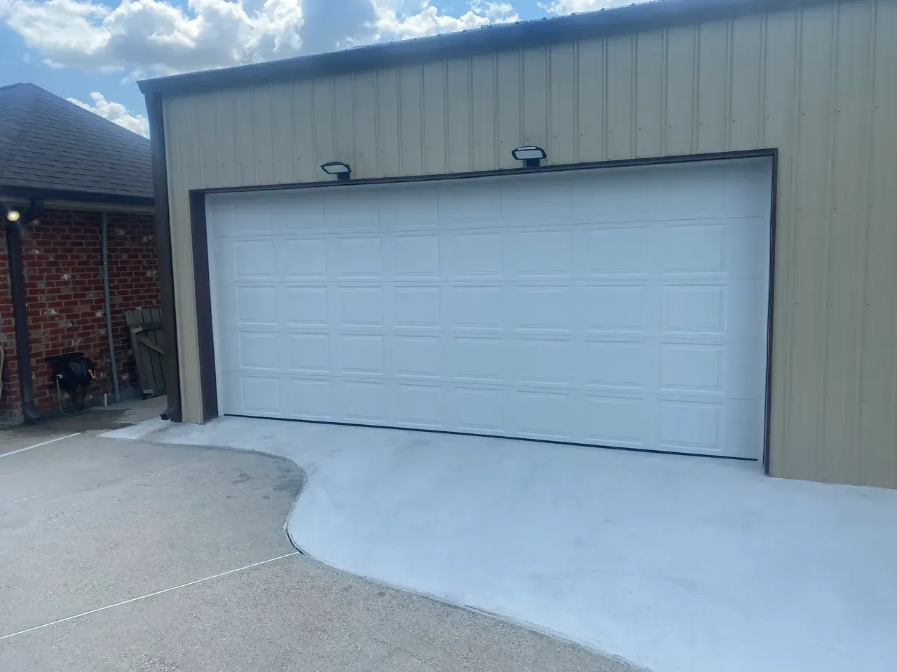 White garage door on a tan metal building with a concrete driveway and brick wall.