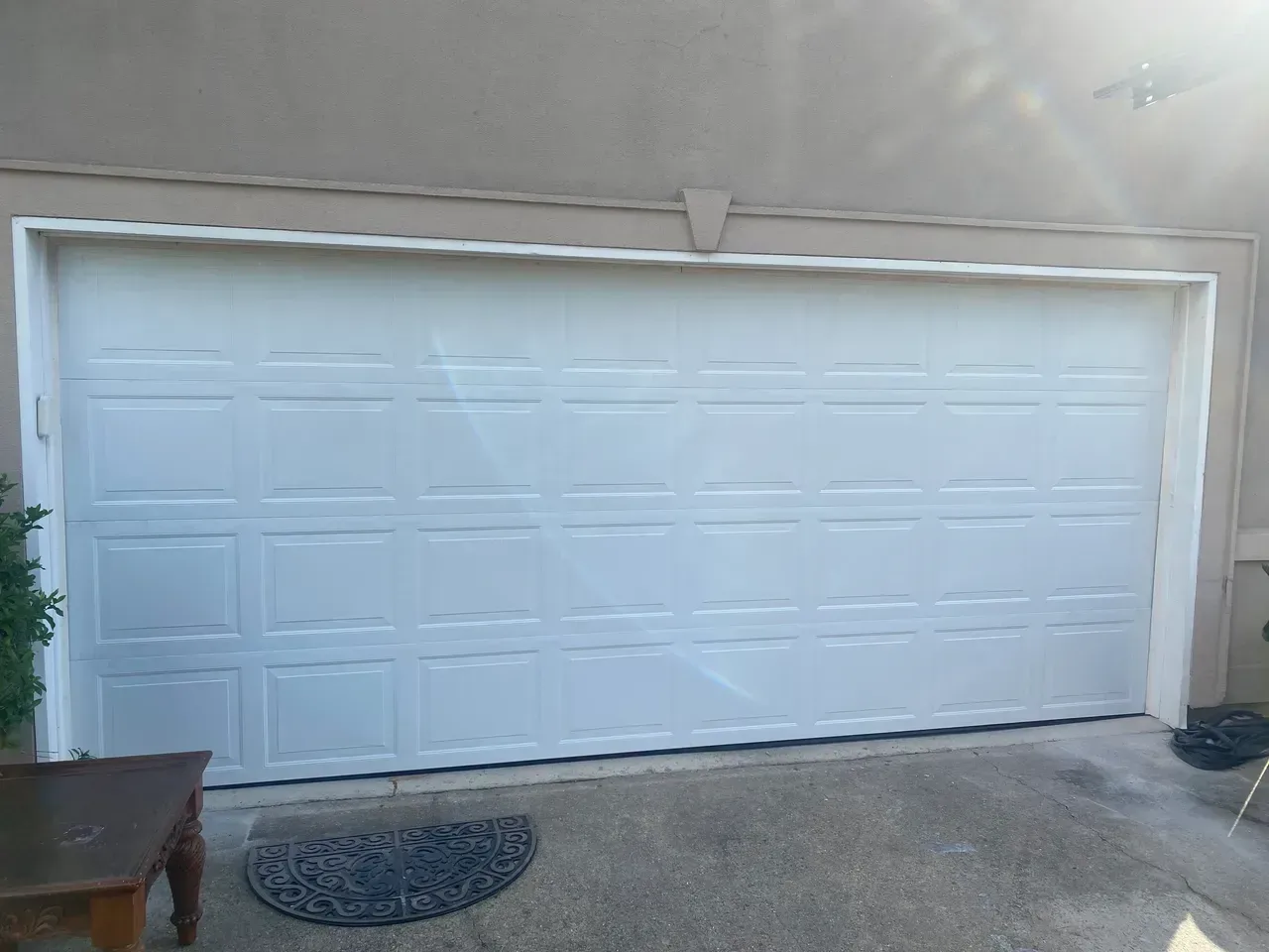 White garage door with rectangular panels on a light beige stucco exterior. A dark doormat and small table are in front.