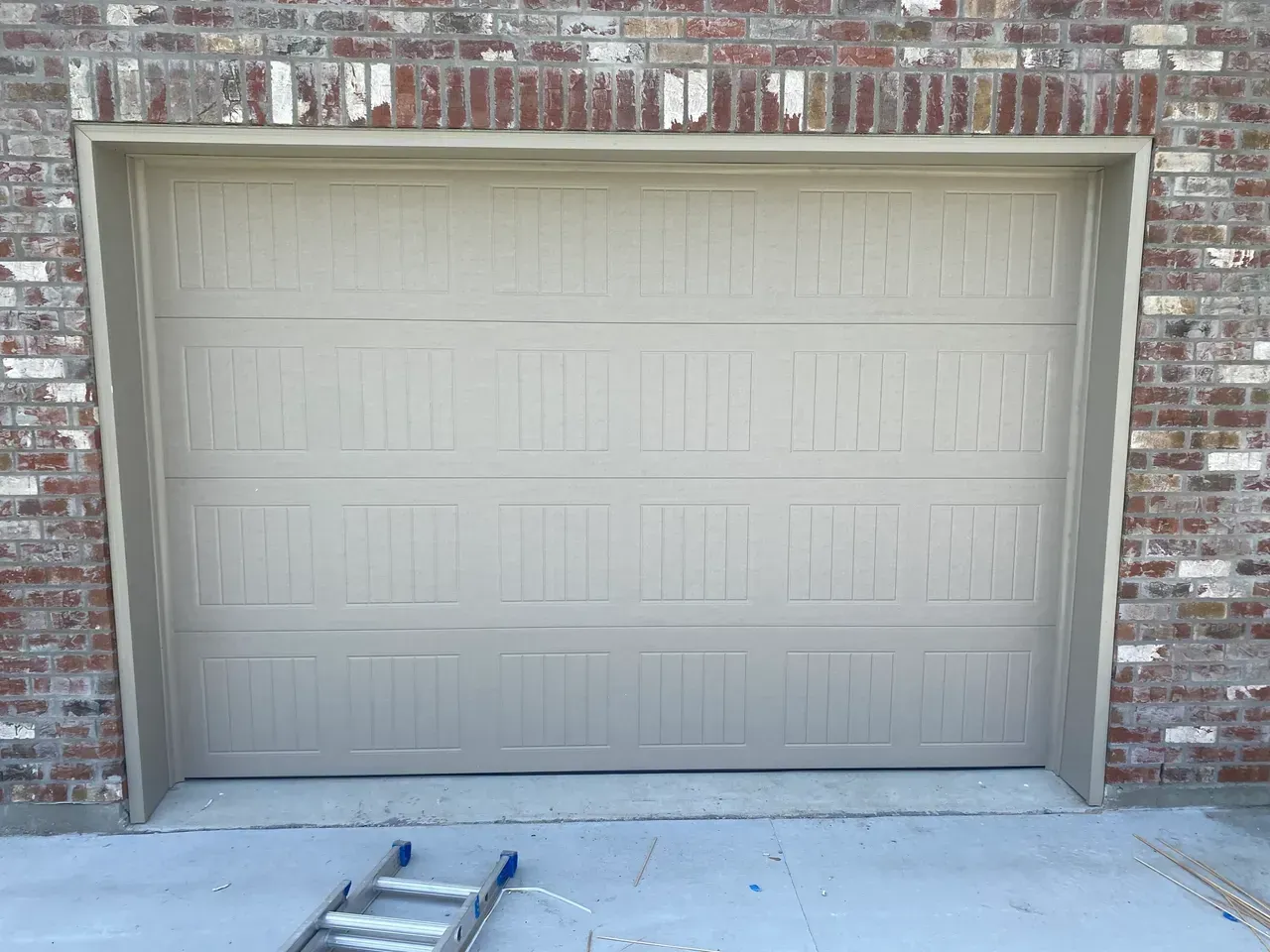 Tan garage door framed by brick and tan trim. A ladder is in front.