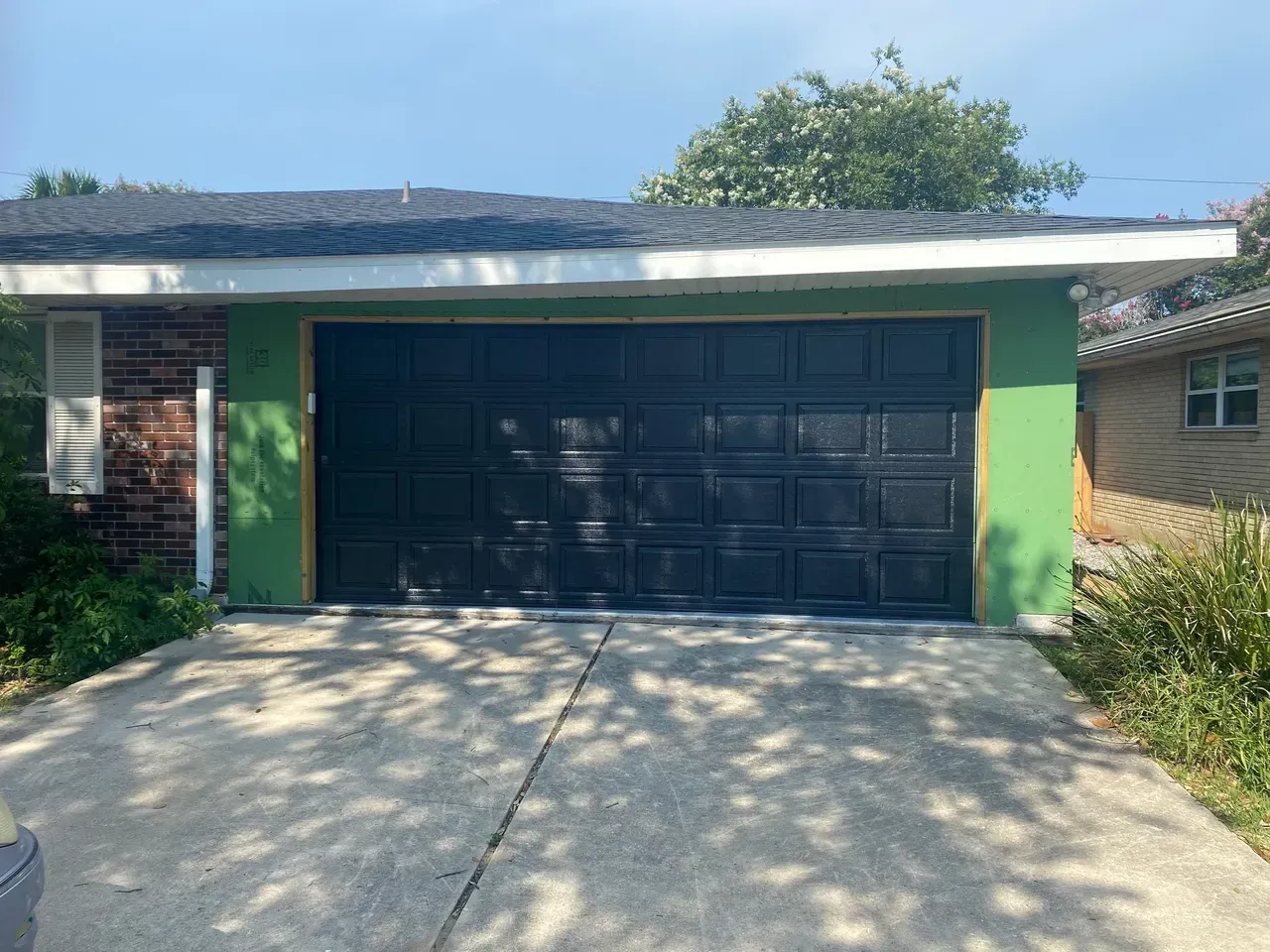 Black garage door on a green-framed garage under a dark roof, with a concrete driveway.