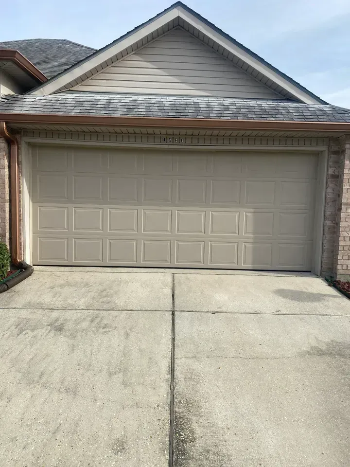 Tan garage door with driveway in front. House with brown trim and roof.