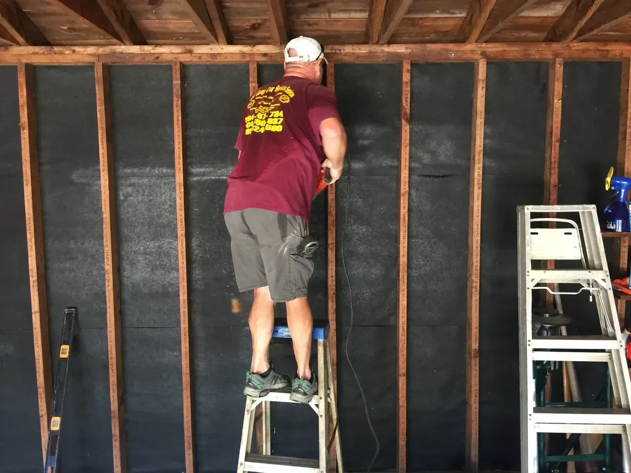 A person on a step ladder cuts something on a wall inside a garage. The wall has black material covering it.