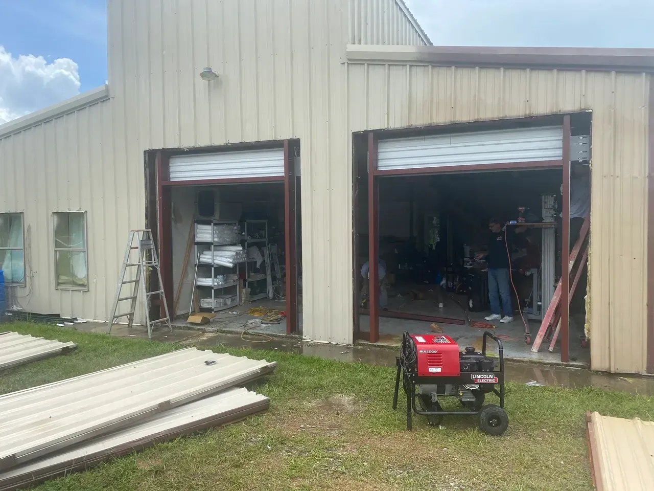 Exterior of a beige metal building with open garage doors. A generator sits in front. Debris and people are visible inside.