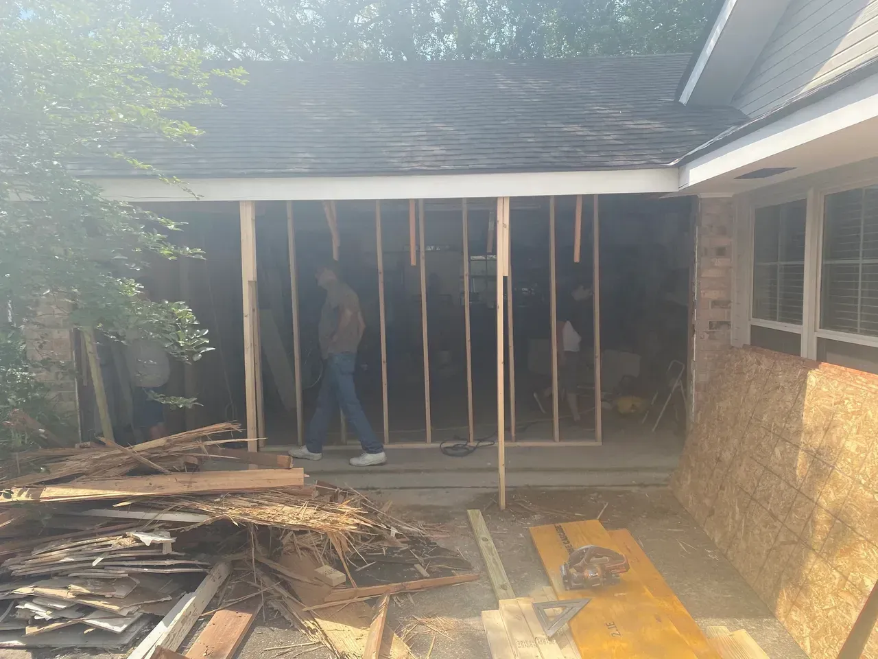 Construction site: Person framed by wood studs, under roof of house, surrounded by debris.