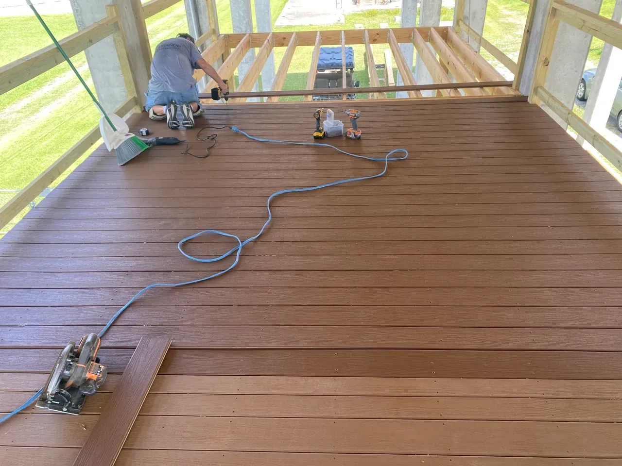 Person installing brown composite deck boards on a wooden frame structure, tools visible.