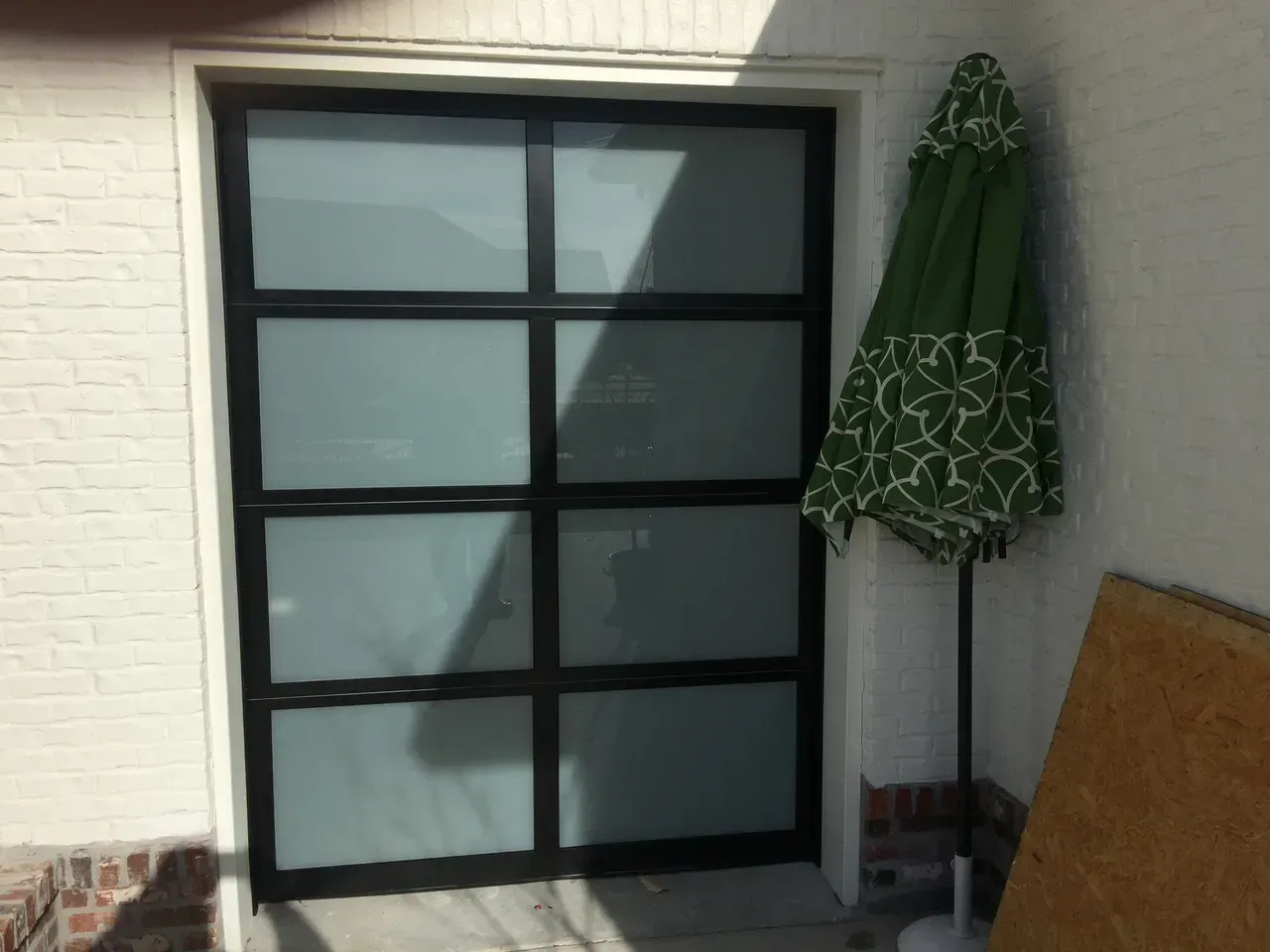 Black framed glass door, white brick wall, green closed patio umbrella, and wooden board.