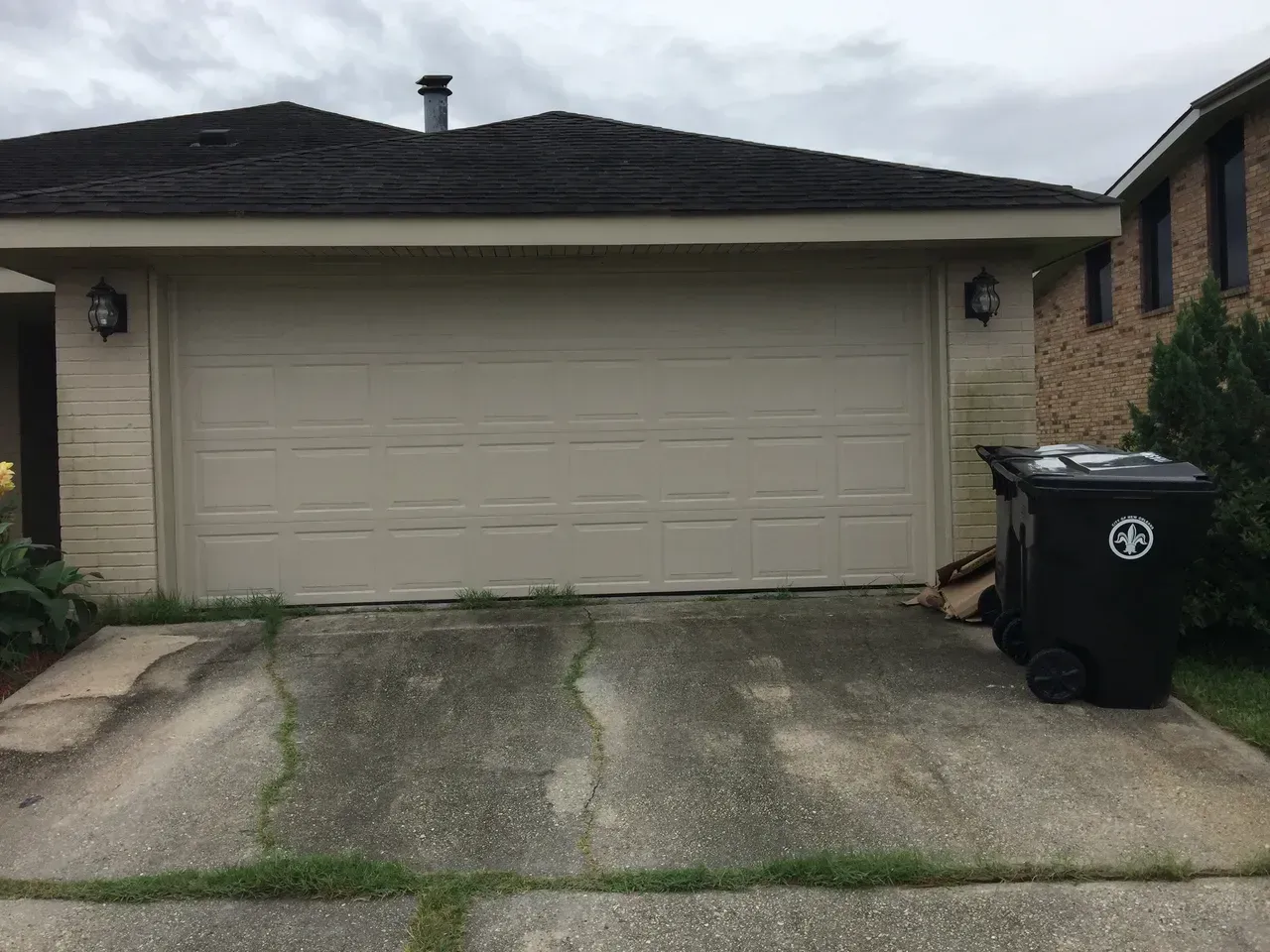Garage with tan door, beige siding, cracked concrete driveway, and black trash bin.