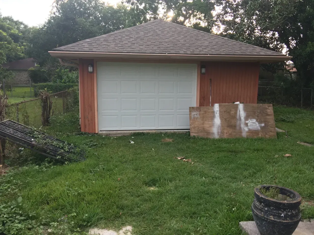 Small garage with a white door, brown roof, and brown siding on a grassy lawn.