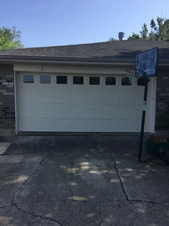 White garage door with six square windows above, basketball hoop to the right, and cracked driveway.