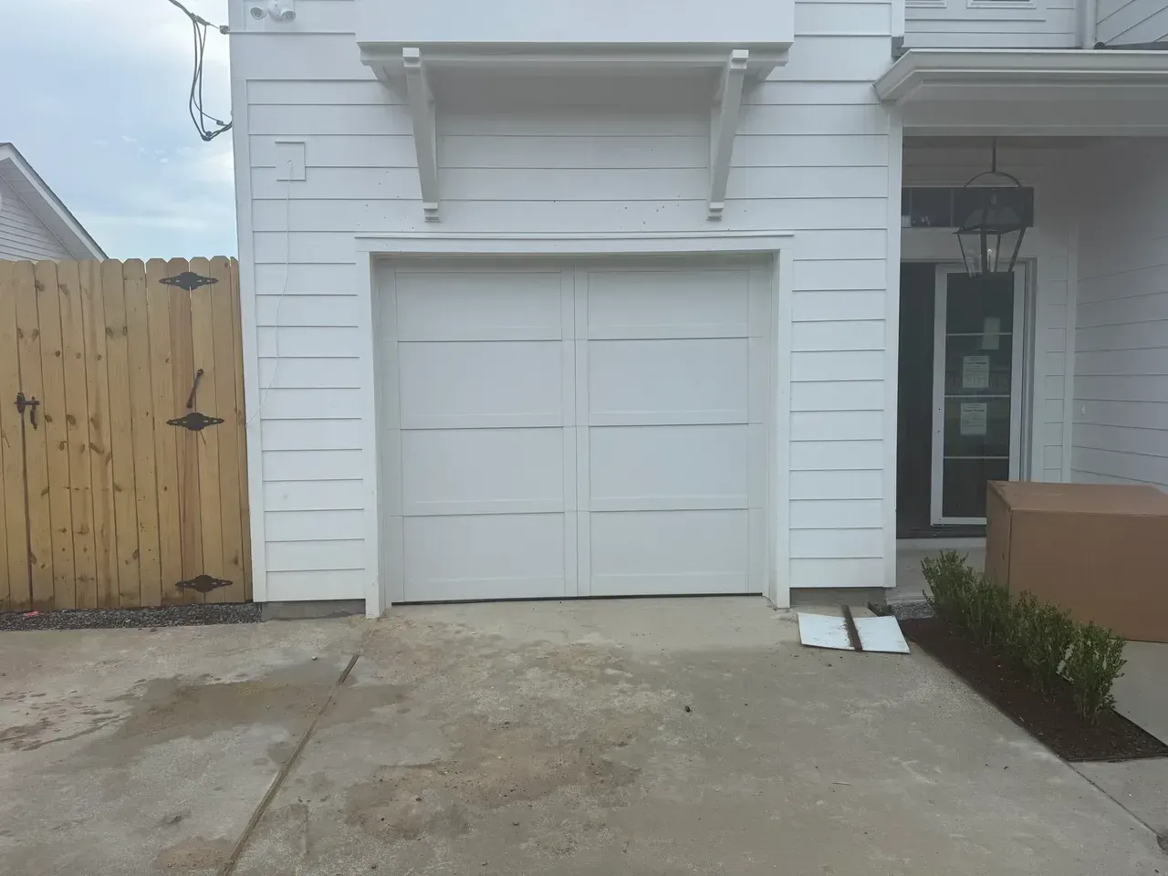 White garage door with a concrete driveway. A wooden fence is on the left, and a door is on the right.