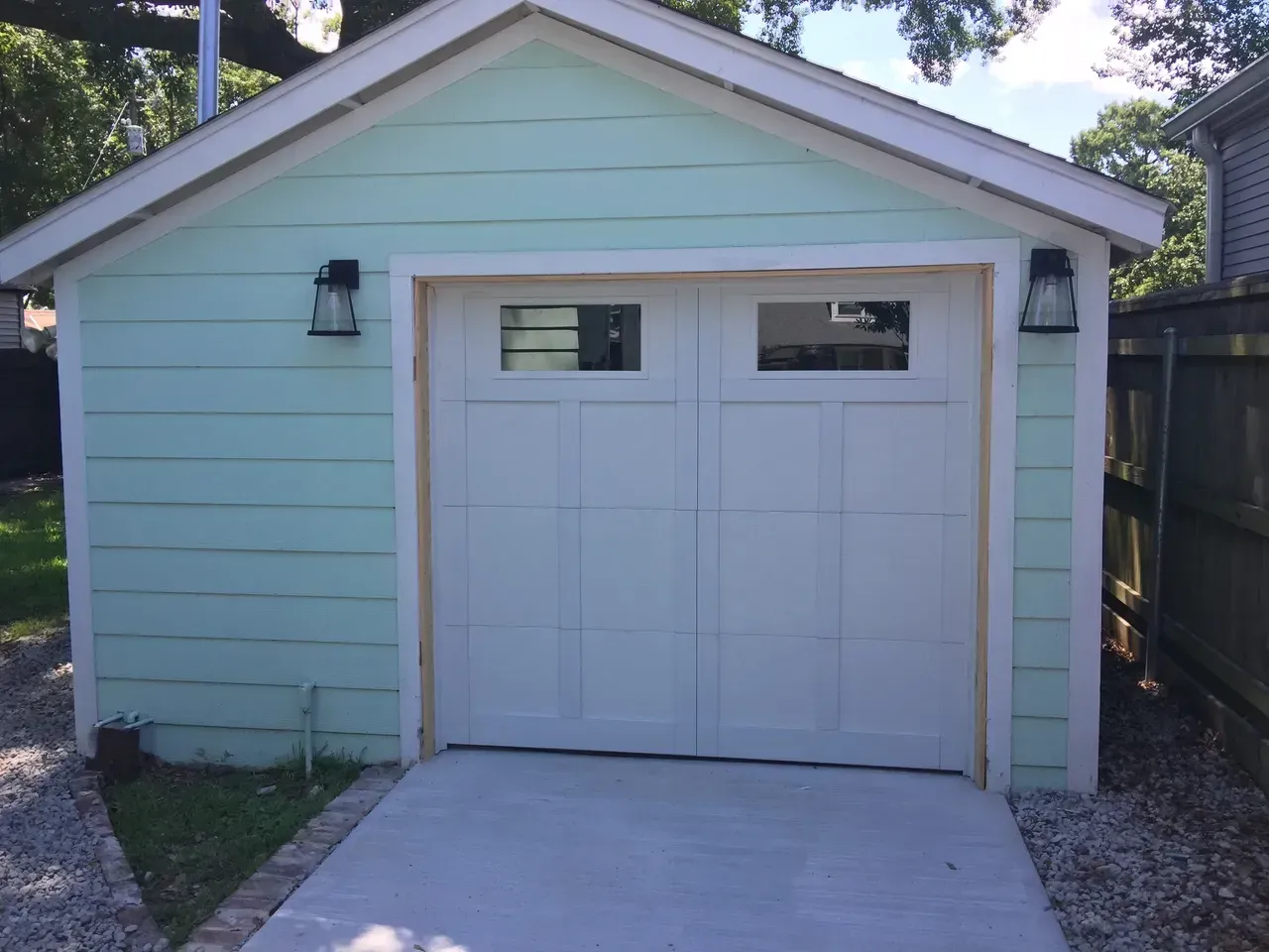 A light-green garage with a white door, two small windows, and black sconces. Concrete driveway.