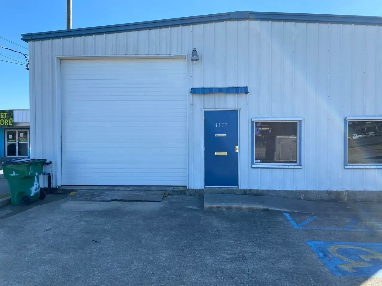 Exterior of a white industrial building with a closed garage door and a blue door. A green trash can sits on the left.