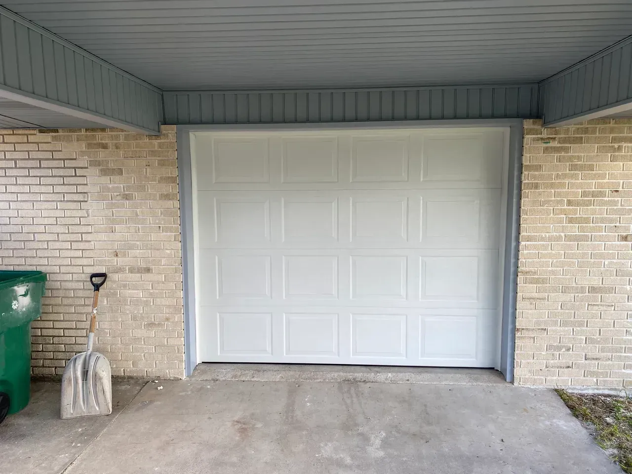White garage door in a brick building with a green trash can and shovel.