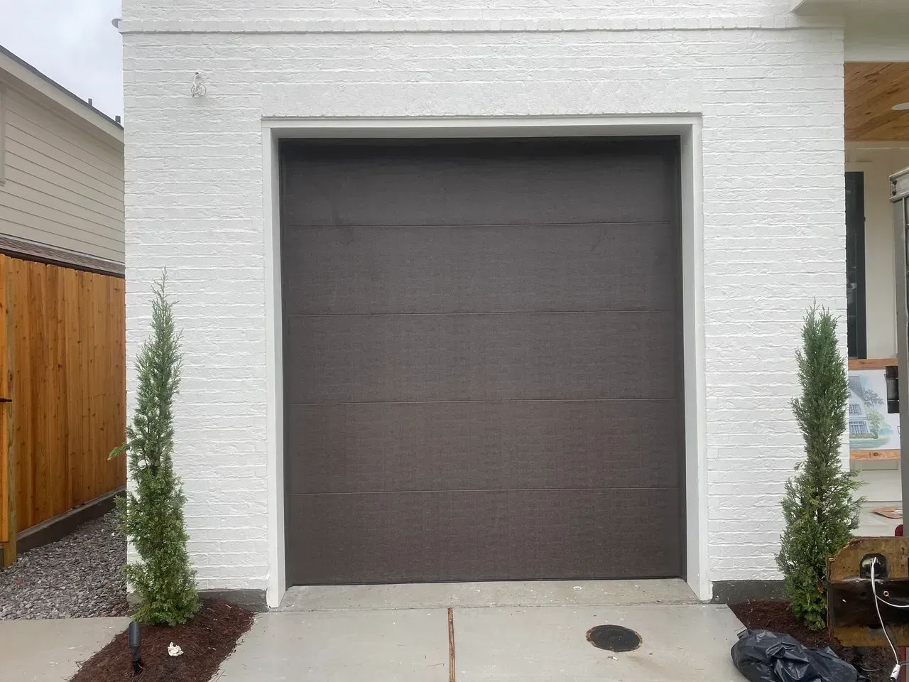 Brown garage door on a white textured building, with small green trees on each side.