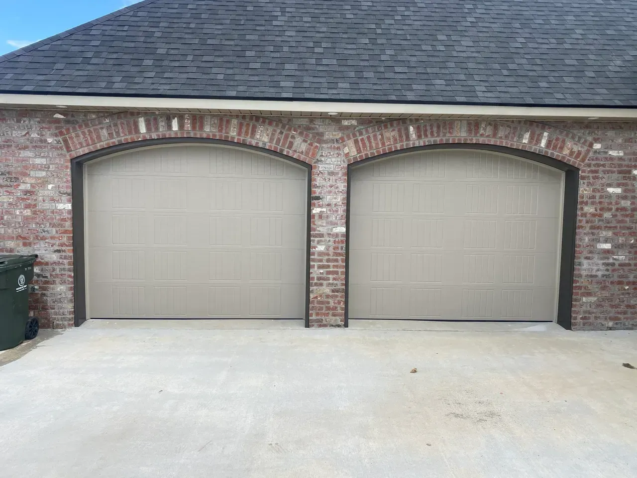 Two tan garage doors under brick arches on a building with a gray roof and concrete driveway.