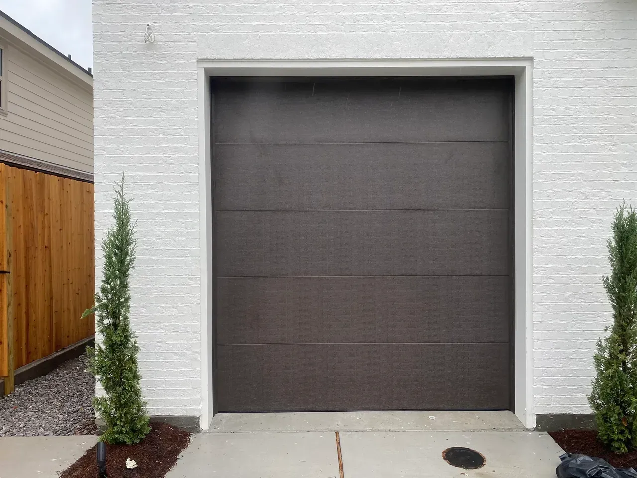 Brown garage door on a white textured wall with two green plants in front of it.