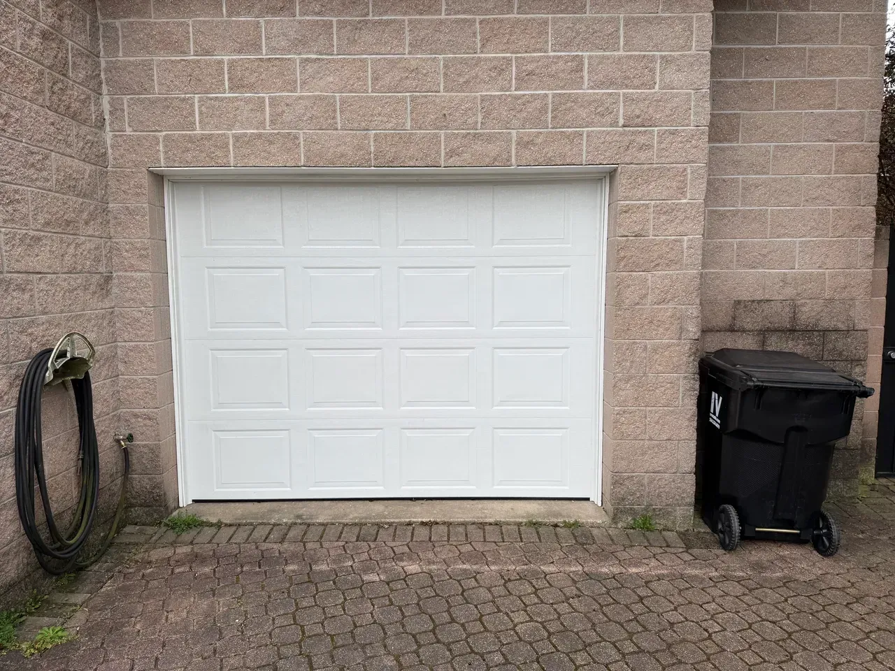 White garage door set in a brick building. A coiled black hose and black trash bin are next to it.