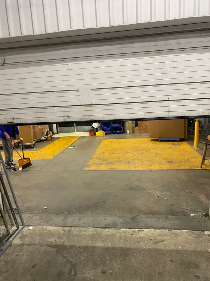 Warehouse loading dock, partially open door, person sweeping. Yellow ramp, boxes, blue bins visible.