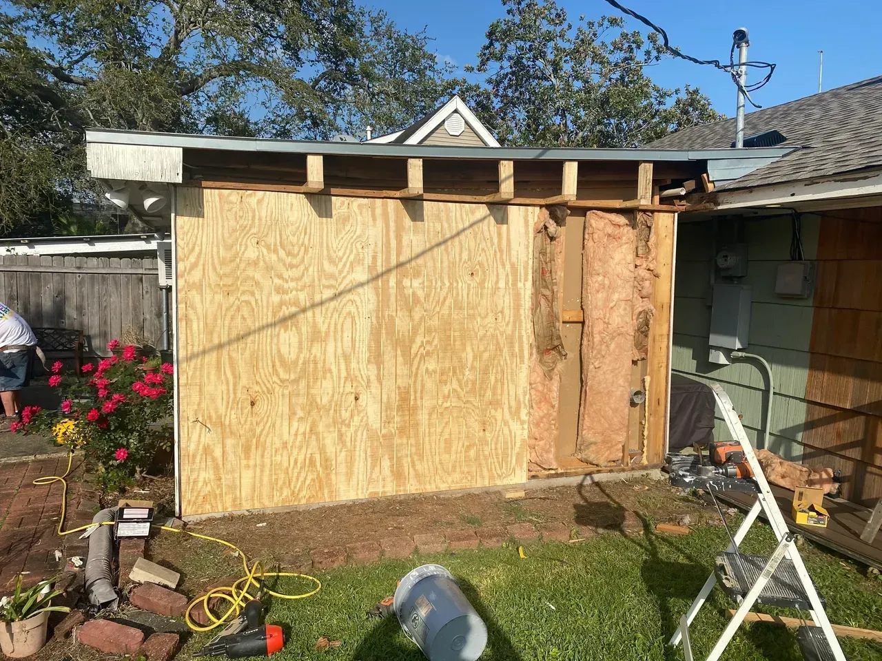 Exterior of a shed under construction; plywood siding, exposed insulation, yard, tools, sunny day.