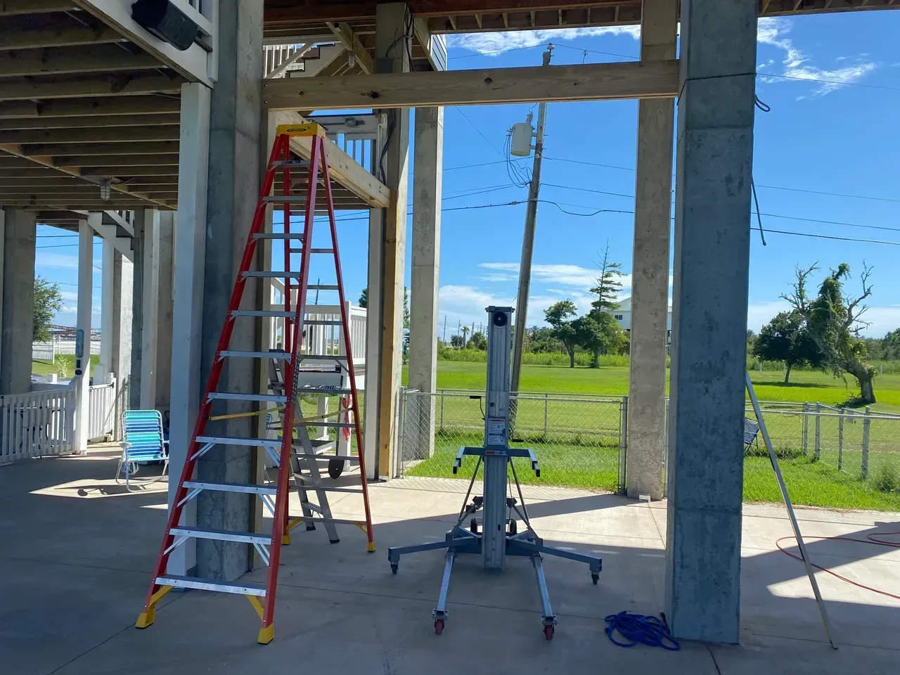 Ladder and lift beneath a building's elevated deck, outdoors with a green field and blue sky in the background.