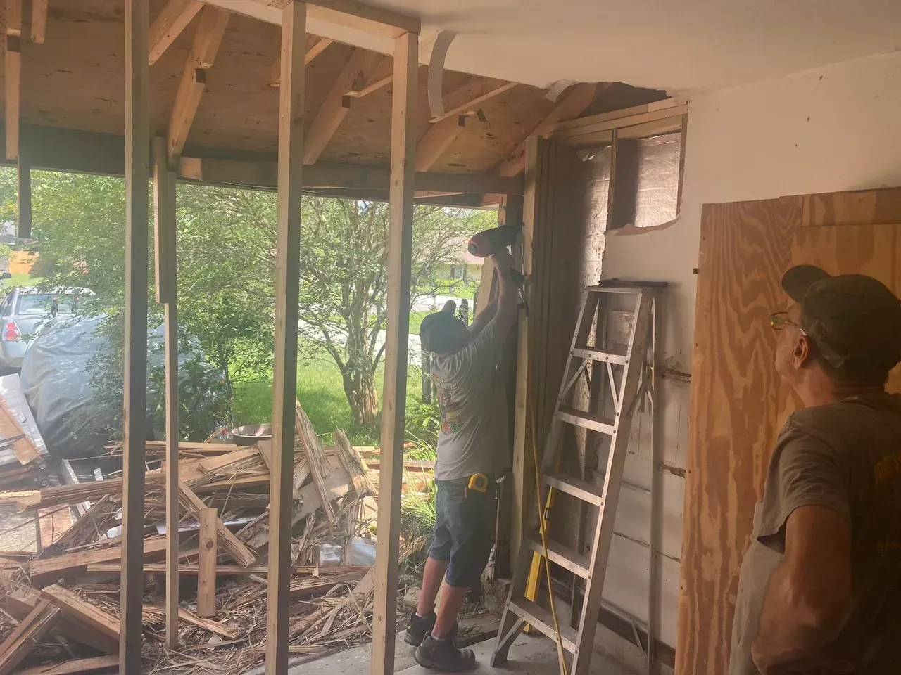Construction workers in a room with exposed framing, using tools to renovate. Ladder and insulation are present.