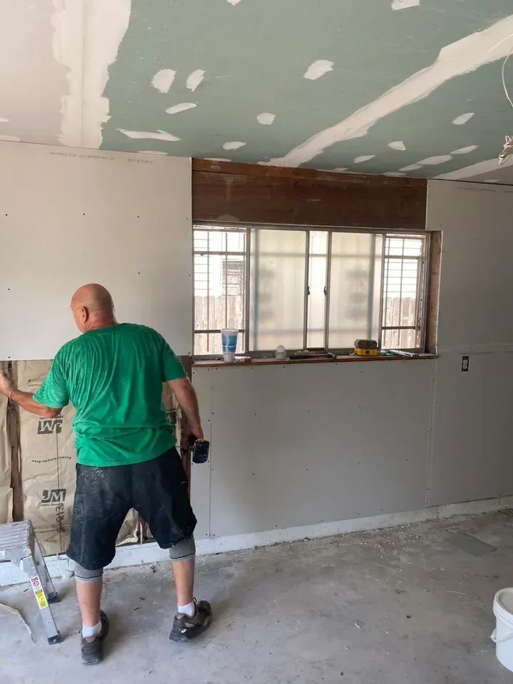 Man installing drywall on a wall with a window, interior construction site.