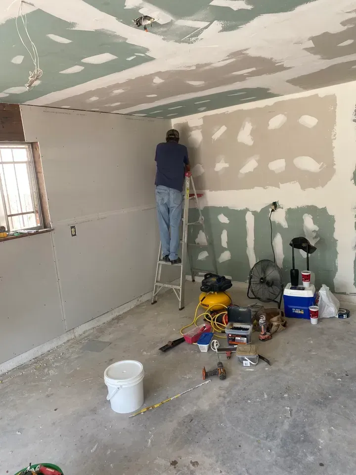 Person on ladder installing drywall in a room with partially finished walls and ceiling. Tools and bucket on the floor.