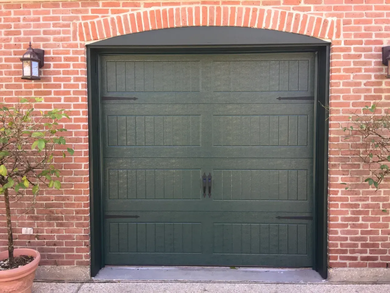 Green garage door in a brick archway. Flanked by brick walls and small trees.