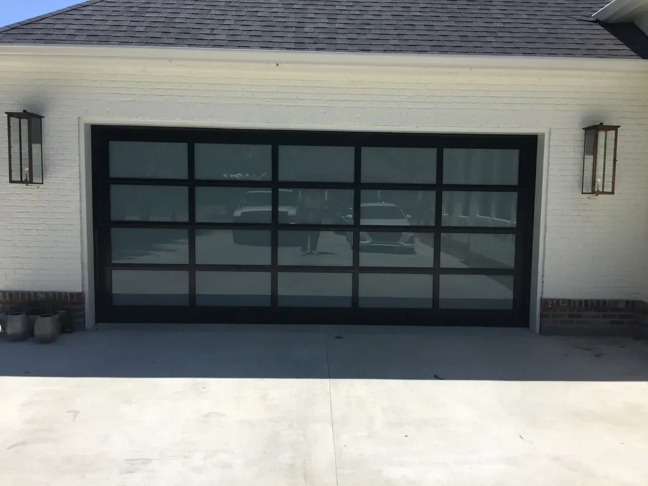 Black-framed glass garage door reflects vehicles and other objects. White brick building with two sconces.